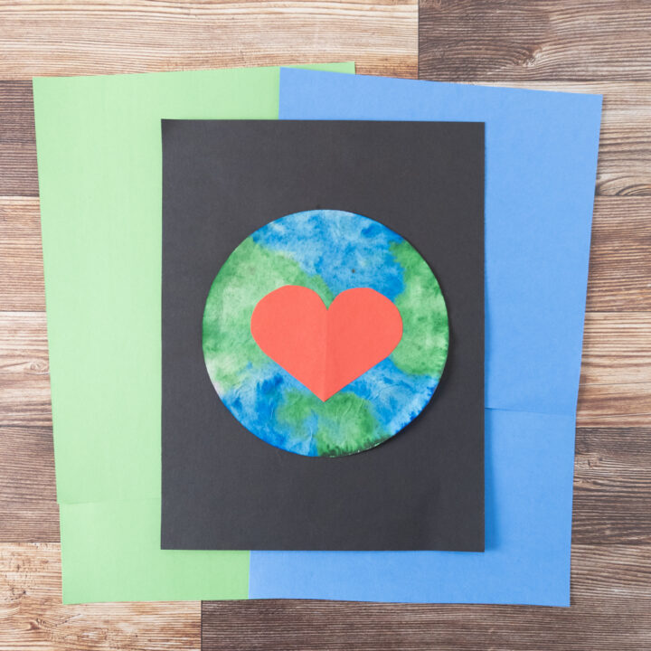 Completed Earth Day coffee filter art project. Green and blue colored filter with big red paper heart glued to the middle. Planet is glued to black paper and laying on top of green and blue papers.