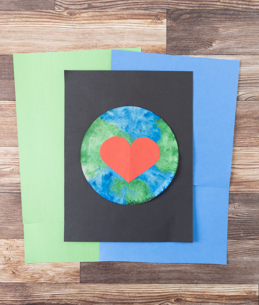 Completed Earth Day coffee filter art project. Green and blue colored filter with big red paper heart glued to the middle. Planet is glued to black paper and laying on top of green and blue papers.