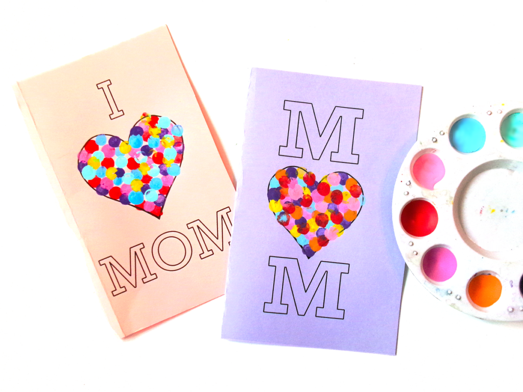 One design is printed on pink paper, and the other on light purple paper. Both have multicolored fingerprint hearts. Next to them is a palette with paints in it.