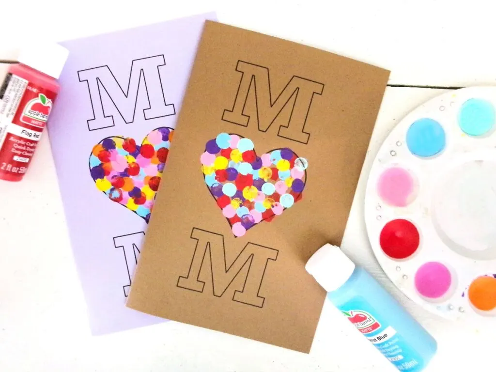 Lavender and gold cardstock used to print out two cards that say MOM vertically with a heart for the "O." Differe colored paint used to make fingerprints in the heart. Red and blue paint bottles and paint palette lay next to cards.