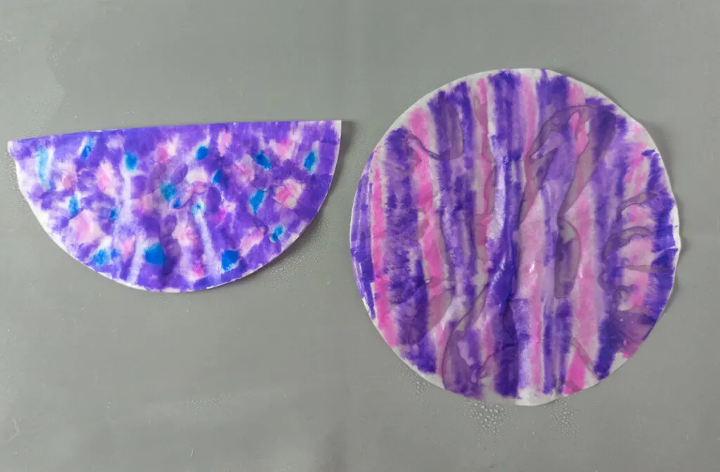 Two round filter papers laying on gray craft mat. Left one is folded in half and colored purple with pink and blue dots. Right one has pink and purple stripes. They are both sprayed with water.