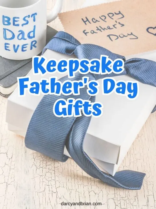 Blue text outlined with white says Keepsake Father's Day Gifts in the center over a background with a white gift box wrapped with a blue tie. A mug with 