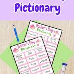 Two pages of Pictionary word cards overlapping on a green paper on a desk. Marker and sand timer laying next to the pages. At the top is dark purple text outlined in white that says Mother's Day Pictionary on a light purple background.