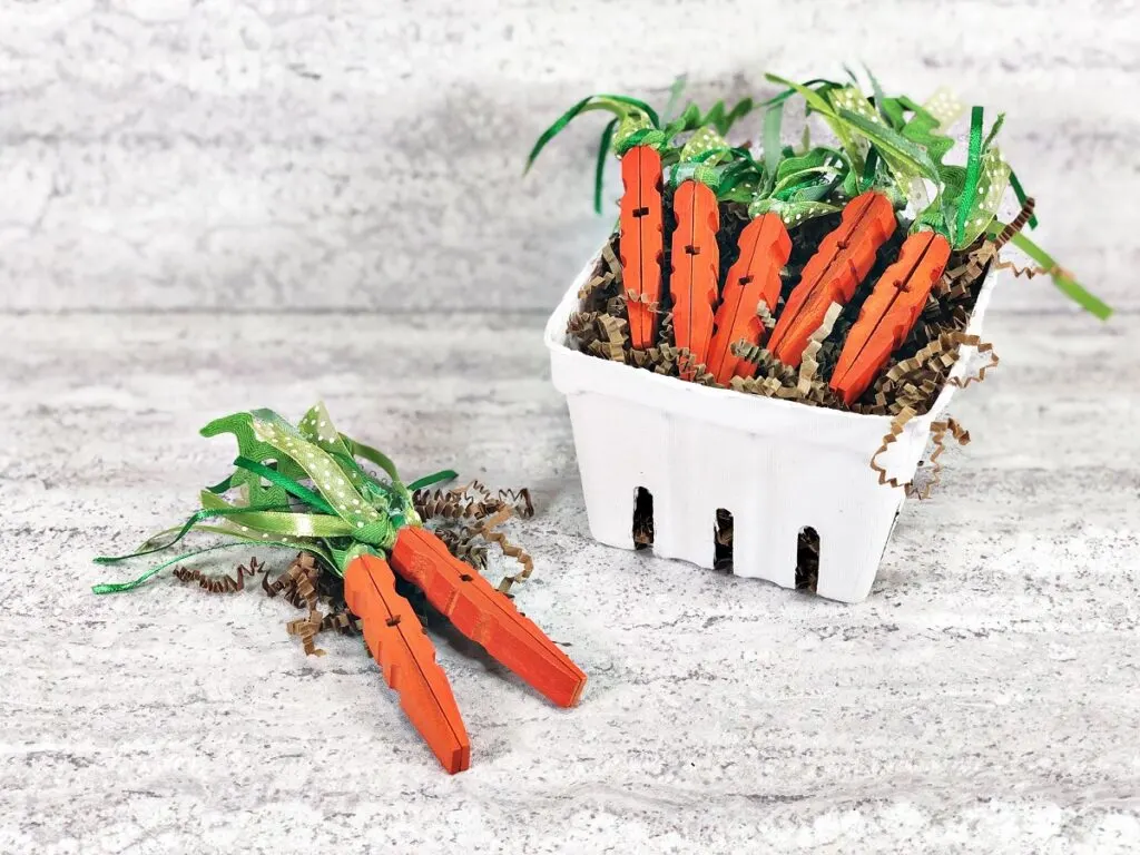 5 completed clothespin carrots laying on top of brown paper shred in a small white box made to look like a vegetable carton. Two more carrots lay on the table next to the box.