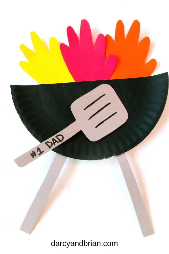 Finished craft with grill made out of black paper plate, paper handprint flames, and gray paper spatula with #1 Dad written on the handle.