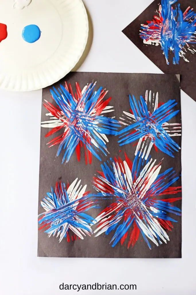 Top down view of completed fork firework painting. Red, white, and blue paint layered on top of each other on black paper to look like fireworks in the sky.