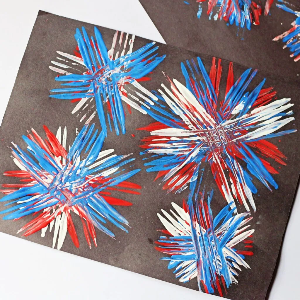 Red, white, and blue paint applied to black paper with a fork creating colorful bursts.