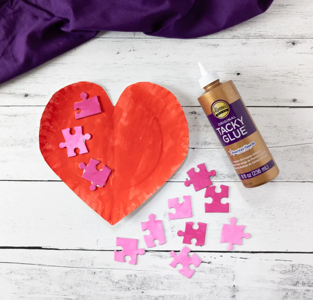 Red painted heart-shaped paper plate with three pink painted puzzle pieces glued to it. Assorted painted jigsaw pieces lay next to it and a bottle of tacky glue.