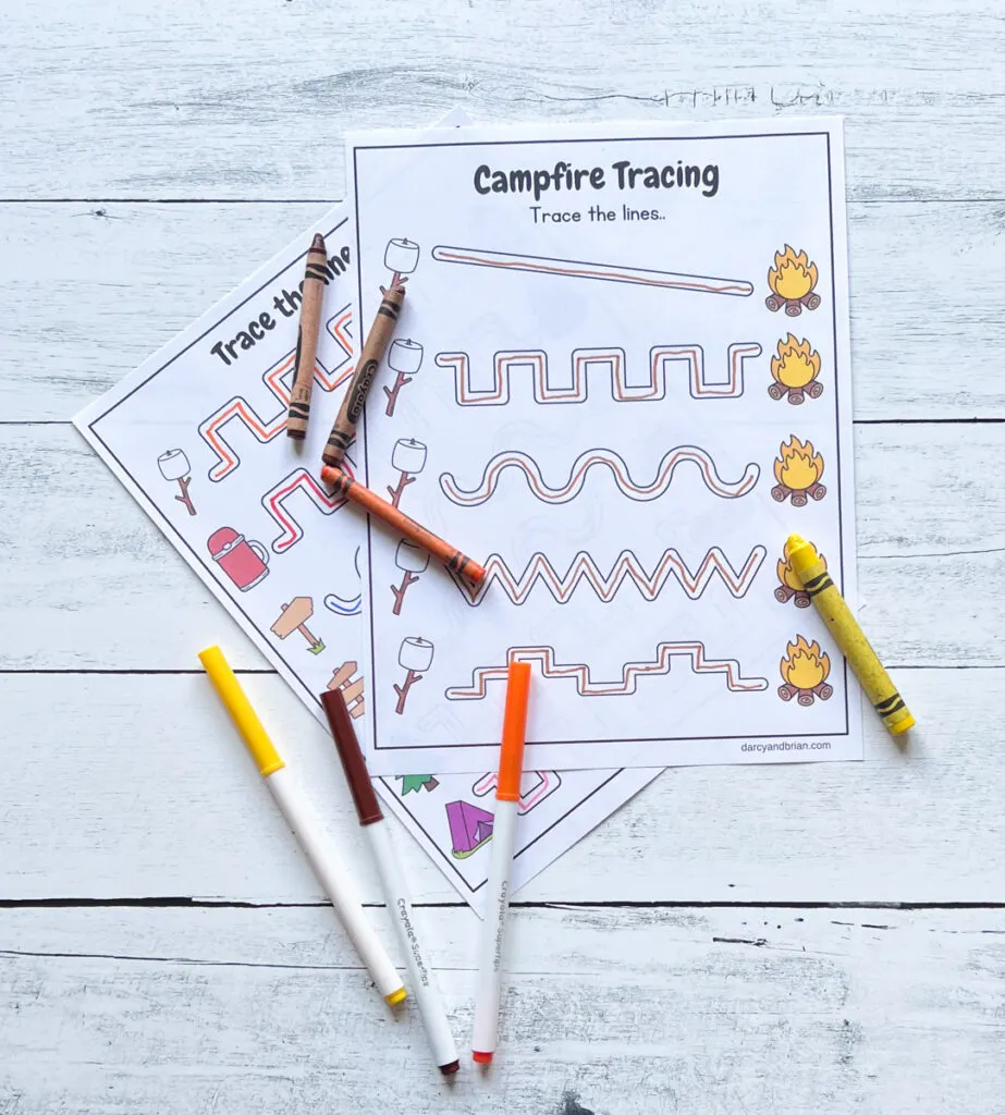Line paths for pencil control in a variety of styles between campfire images. Markers and crayons are lying across the papers.