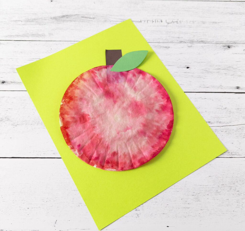 Top down angled view of finished craft to make an apple using a coffee filter. The red apple is laying on bright green paper.