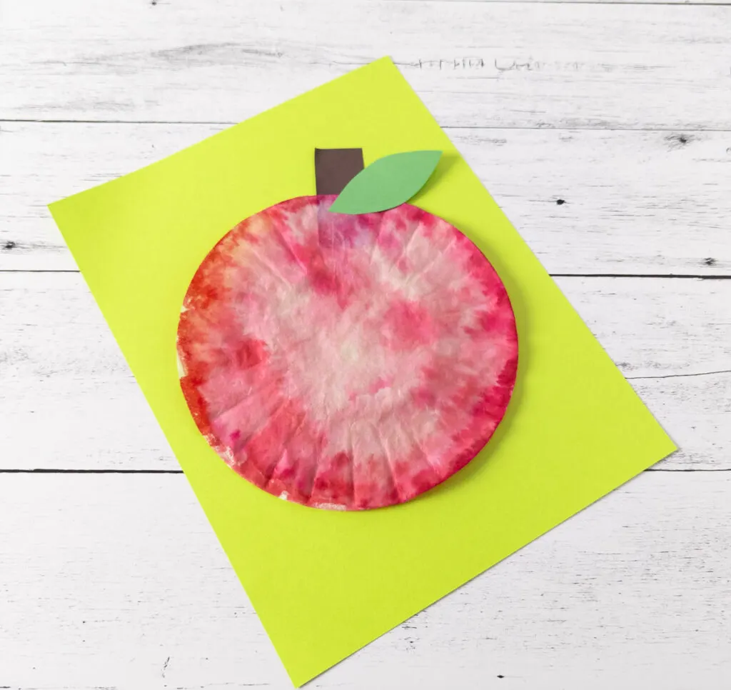 Top down angled view of finished craft to make an apple using a coffee filter. The red apple is laying on bright green paper.
