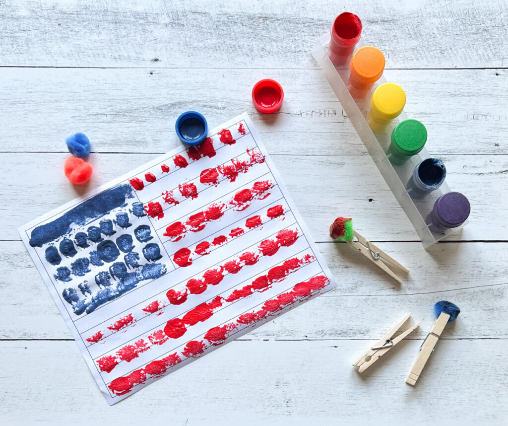 Overhead view of completed flag covered with blue and red dabs of paint. Fuzzy balls clipped in clothespins lay next to paint.