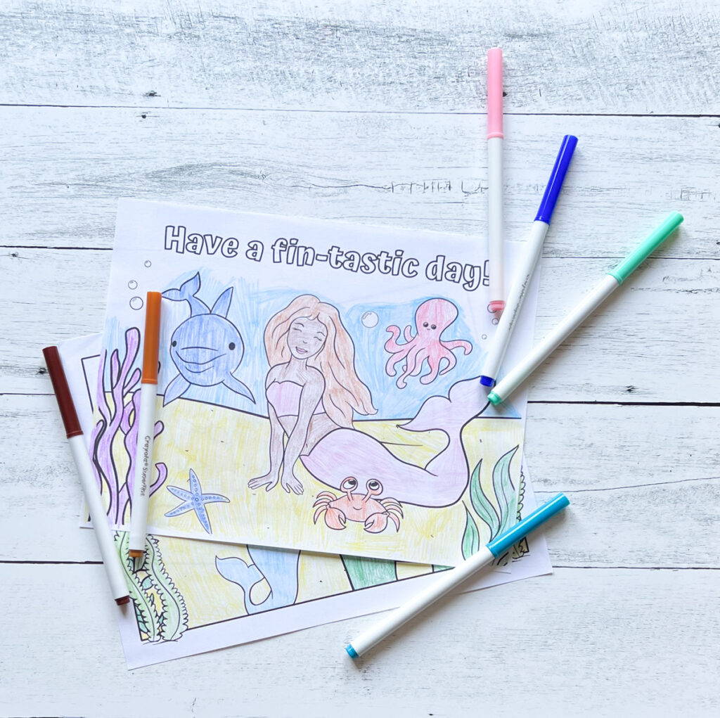 Paper with mermaid ocean scene colored in. Text at top of page says Have a fin-tastic day!. Markers lay around the page.